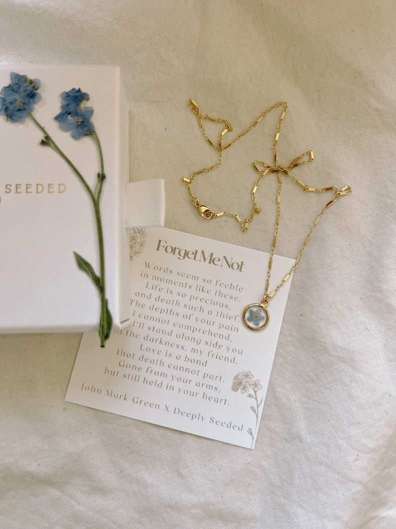May include: Gold necklace featuring a forget-me-not flower pressed within a glass pendant. The necklace is displayed on a white card with the text "Forget Me Not" and a poem about love and loss.