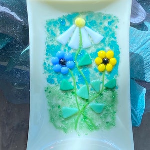 May include: A white rectangular dish with a floral design made of glass. The design features a white daisy, a blue flower, and a yellow flower with green leaves.