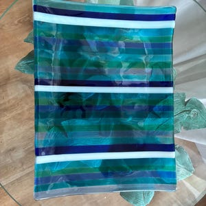 May include: A rectangular fused glass tray featuring a repeating horizontal striped pattern in shades of teal, turquoise, dark blue, and white. The glass surface is smooth and glossy, with a translucent quality that allows light to pass through.