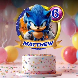 Peut inclure: Un gâteau blanc glacé avec des vermicelles colorés, surmonté d'un décor de gâteau Sonic le Hérisson. Le décor représente le personnage, le nom "MATTHEW" et le chiffre 6. Des ballons de différentes couleurs sont en arrière-plan.