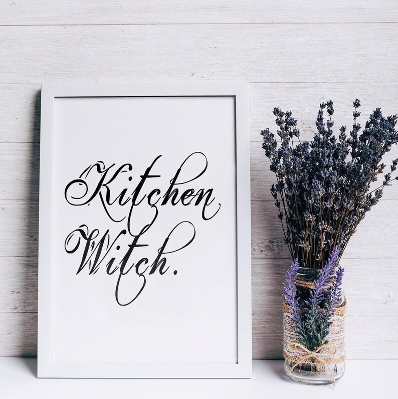 Puede incluir: Impresi&oacute;n enmarcada en blanco con caligraf&iacute;a negra que dice "Kitchen Witch" sobre un fondo de madera blanca. Un ramo de lavanda est&aacute; en un jarr&oacute;n de vidrio transparente con una cinta de arpillera.