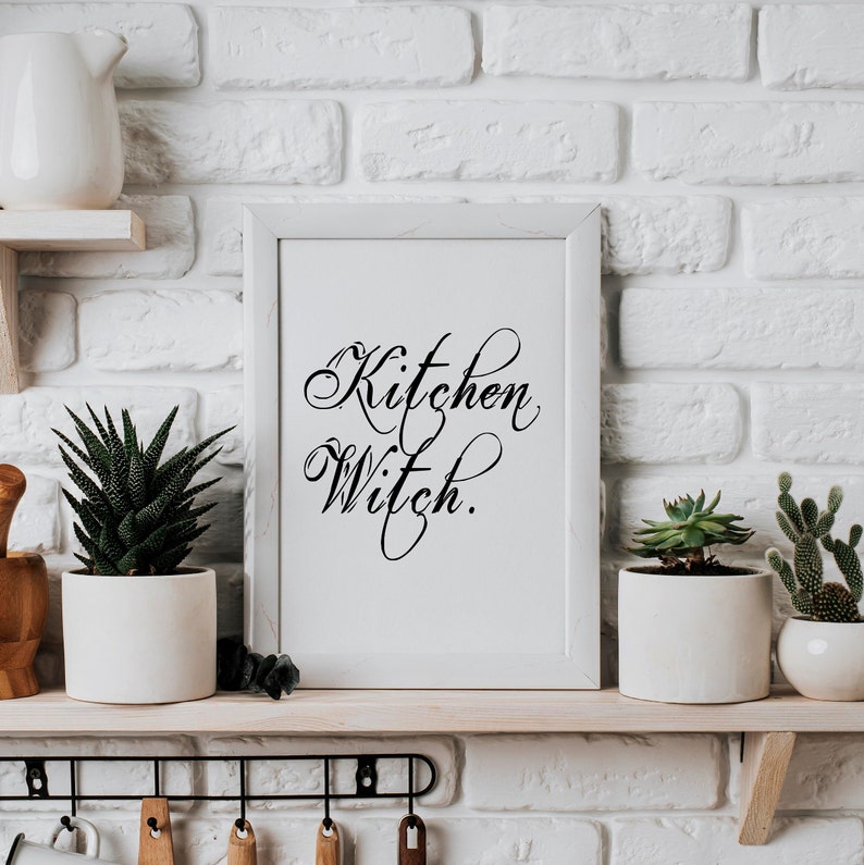 Puede incluir: Impresi&oacute;n enmarcada en blanco con caligraf&iacute;a negra que dice "Kitchen Witch." La impresi&oacute;n se muestra en un estante blanco con una pared de ladrillo blanco en el fondo.
