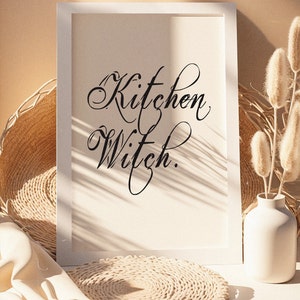 Puede incluir: Impresi&oacute;n enmarcada en blanco con el texto "Kitchen Witch." La impresi&oacute;n est&aacute; sobre una superficie blanca con una estera de paja tejida y un jarr&oacute;n blanco con flores secas beige.