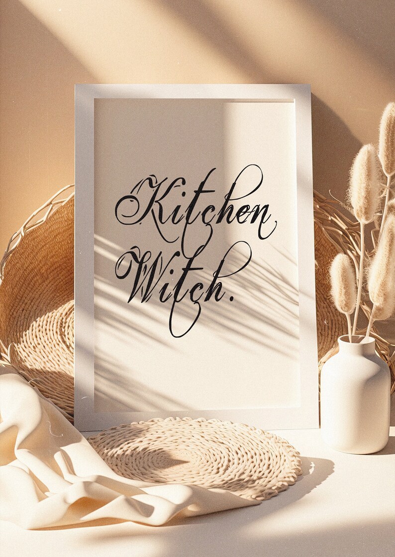 Puede incluir: Impresi&oacute;n enmarcada en blanco con el texto "Kitchen Witch." La impresi&oacute;n est&aacute; sobre una superficie blanca con una estera de paja tejida y un jarr&oacute;n blanco con flores secas beige.