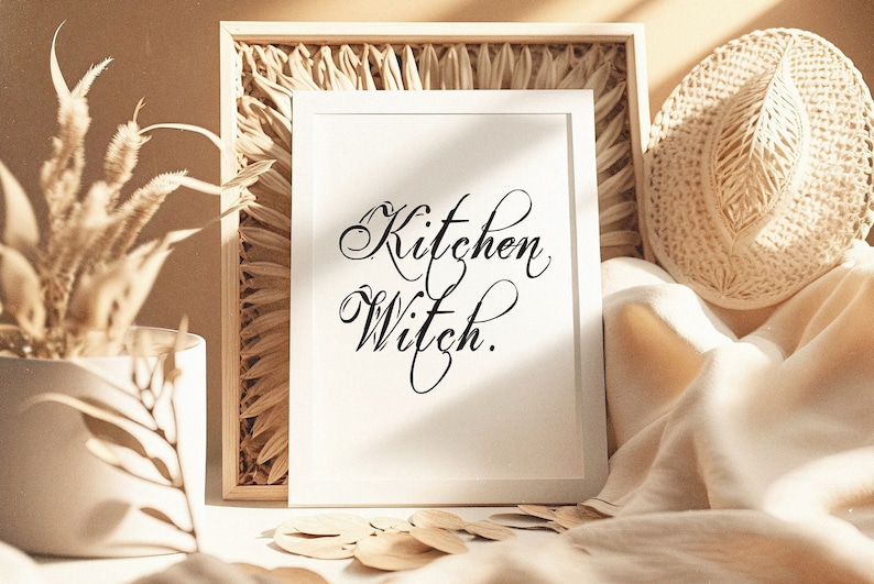 Puede incluir: Una impresi&oacute;n enmarcada en blanco con el texto "Kitchen Witch." en escritura cursiva negra. La impresi&oacute;n est&aacute; sobre una mesa con una cesta tejida, un sombrero de paja y plantas secas.
