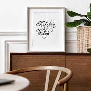 Puede incluir: Una impresi&oacute;n enmarcada con el texto "Kitchen Witch" en una fuente de escritura decorativa. La impresi&oacute;n est&aacute; colgada en una pared sobre un armario de madera.