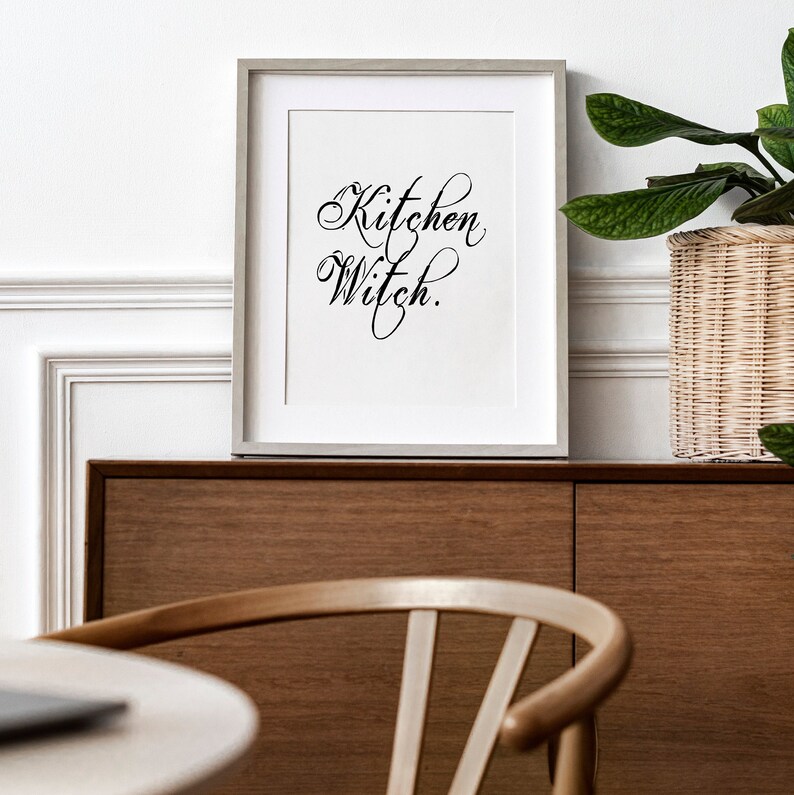 Puede incluir: Una impresi&oacute;n enmarcada con el texto "Kitchen Witch" en una fuente de escritura decorativa. La impresi&oacute;n est&aacute; colgada en una pared sobre un armario de madera.