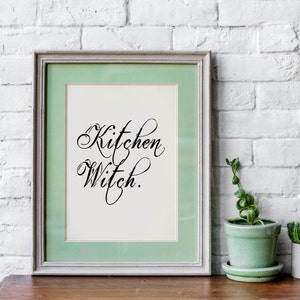 Puede incluir: Una impresi&oacute;n enmarcada con un passe-partout verde claro y un fondo blanco. La impresi&oacute;n presenta el texto "Kitchen Witch" en una fuente de escritura decorativa.