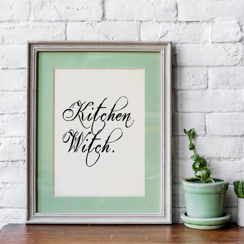 Puede incluir: Una impresi&oacute;n enmarcada con un passe-partout verde claro y un fondo blanco. La impresi&oacute;n presenta el texto "Kitchen Witch" en una fuente de escritura decorativa.