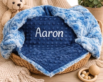 May include: A blue baby blanket with the name "Aaron" embroidered in white. The blanket is inside a woven basket and has a soft, plush texture. A teddy bear and a wooden toy are also in the image.