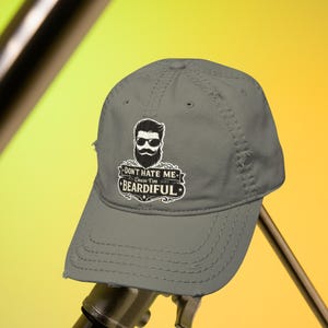 May include: A grey baseball cap with the slogan "DON'T HATE ME Cause I'm BEARDIFUL" and a graphic of a bearded man wearing sunglasses. The cap has a curved peak and an adjustable strap.