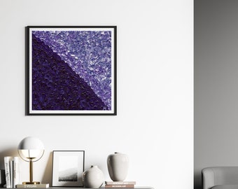 May include: A framed abstract painting with a diagonal line separating two shades of purple. The painting is hanging on a white wall above a grey floating shelf with books, vases, and a lamp.
