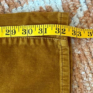 以下が含まれることがあります： マスタード色のコーデュロイ生地に、黄色のメジャーがかけられています。メジャーは71〜86cmの測定値を示しています。生地はリブ編みのテクスチャです。