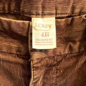 以下が含まれることがあります： 茶色のコーデュロイパンツのクローズアップ。ラベルには「J.CREW」と「4R FAVORITE FIT」と生地の詳細が記載されています。パンツは綿98％、スパンデックス2％で作られています。