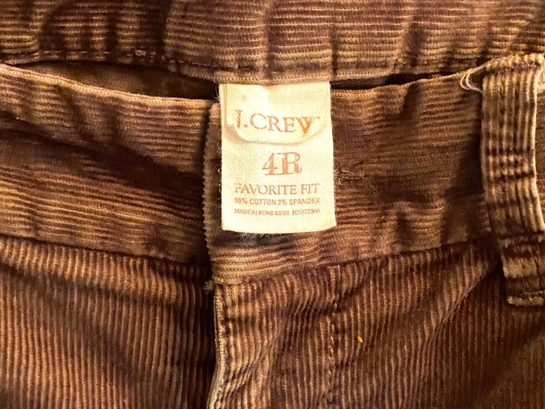以下が含まれることがあります： 茶色のコーデュロイパンツのクローズアップ。ラベルには「J.CREW」と「4R FAVORITE FIT」と生地の詳細が記載されています。パンツは綿98％、スパンデックス2％で作られています。