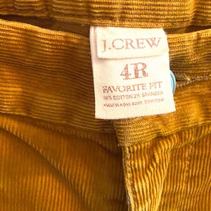 以下が含まれることがあります： マスタードイエローのコーデュロイパンツのクローズアップ。タグには「J.CREW」と「4R FAVORITE FIT」と記載されています。生地は綿98％、スパンデックス2％です。パンツは香港製です。