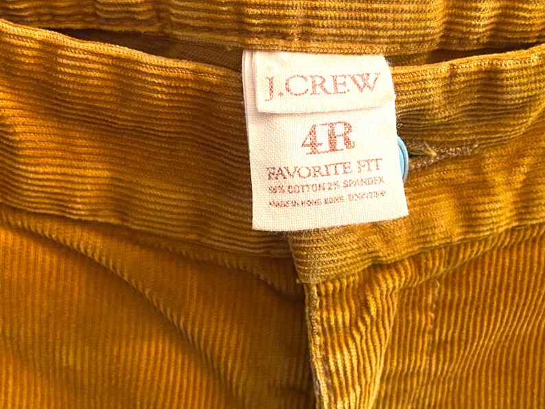 以下が含まれることがあります： マスタードイエローのコーデュロイパンツのクローズアップ。タグには「J.CREW」と「4R FAVORITE FIT」と記載されています。生地は綿98％、スパンデックス2％です。パンツは香港製です。