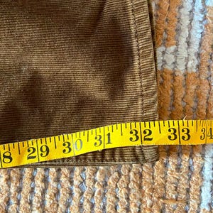 以下が含まれることがあります： 黄色のメジャーが付いた茶色のコーデュロイ生地のクローズアップ。テープは71〜89cmの測定値を示しています。生地には垂直の畝があり、縫い目が見えます。