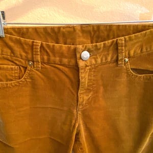 以下が含まれることがあります： マスタード色のコーデュロイパンツのクローズアップ。 パンツは、ボタン留めと金属製のジッパーを備えたクラシックな5ポケットデザインです。 コーデュロイ生地はリブ編みのテクスチャです。 パンツはシルバーのハンガーに吊るされています。