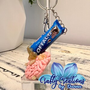 May include: A pink brain-shaped keychain with a blue and white Offalo candy bar keychain charm. The keychain is on a clear acrylic display stand with a blue flower graphic and the text "Crafty Creations by Danna".