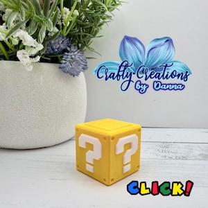 May include: A yellow cube with white question marks on its sides, resembling a video game block. The image includes the text Crafty Creations by Danna and the word CLICK in colorful letters. A potted plant with green and purple foliage is visible.