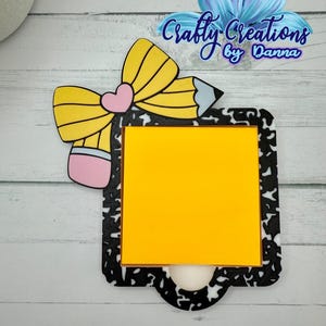 May include: A school-themed notepad holder with a yellow pencil and bow design. The notepad has a black and white speckled border and a bright yellow sticky note. The text "Crafty Creations by Danna" is visible.