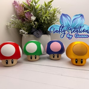 May include: Four mushroom-shaped figures with rounded caps and stems featuring simple facial expressions. The caps are red with white spots, green with white spots, blue with pink spots, and solid yellow. The text Crafty Creations by Danna is visible.
