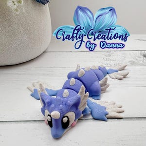 May include: A light blue and white dragon figurine with large eyes and a detailed design. The dragon has a long body with white accents and is lying on a white wooden surface. The image includes the text "Crafty Creations by Danna".