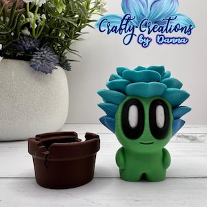 May include: A green and blue 3D printed succulent planter with a brown pot. The planter has a face with two white eyes and a small smile. The text "Crafty Creations by Danna" is visible in the background.