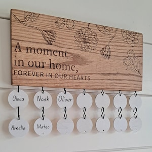 May include: A wooden plaque with the text "A moment in our home, FOREVER IN OUR HEARTS" and floral designs.  The plaque has 10 hanging circles with the names Olivia, Noah, Oliver, Amelia, and Mateo written on some of the circles.
