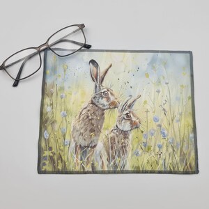 May include: A watercolour painting of two brown rabbits in a field of green grass and blue flowers. The rabbits are looking at each other.