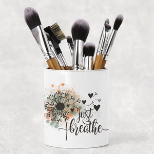 May include: White ceramic makeup brush holder with a dandelion design and the text "Just Breathe". The holder is filled with a variety of makeup brushes.