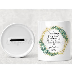 May include: White ceramic money box with a slot on the top. The front of the box has a gold geometric frame with green eucalyptus leaves and the text "Wedding Day Fund Paul & Emma 12th September 2022".