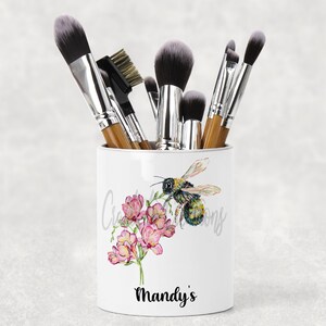 May include: White ceramic makeup brush holder with a bee and pink flowers design. The holder has the text "Mandy's" printed on the bottom.