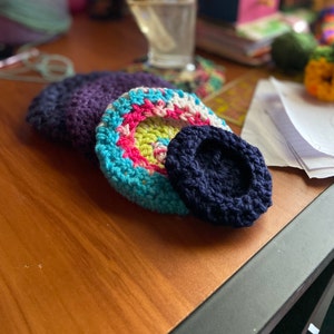 May include: A collection of handmade crochet items in various sizes and colors. The items include a dark purple, a multi-colored piece with blue, pink, and yellow, and a small navy blue piece. The items are arranged on a wooden surface.