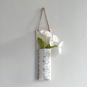 May include: White ceramic wall vase with a blue and white floral pattern. The vase is hanging from a string and has white flowers inside.