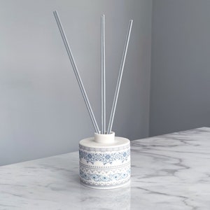 May include: A white ceramic diffuser bottle with a blue and grey patterned design. The bottle has three grey diffuser sticks inserted into the top.
