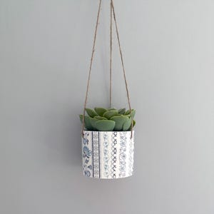 May include: A small, white ceramic planter with a blue and white patterned design hangs from a twine cord. The planter contains a green succulent plant.