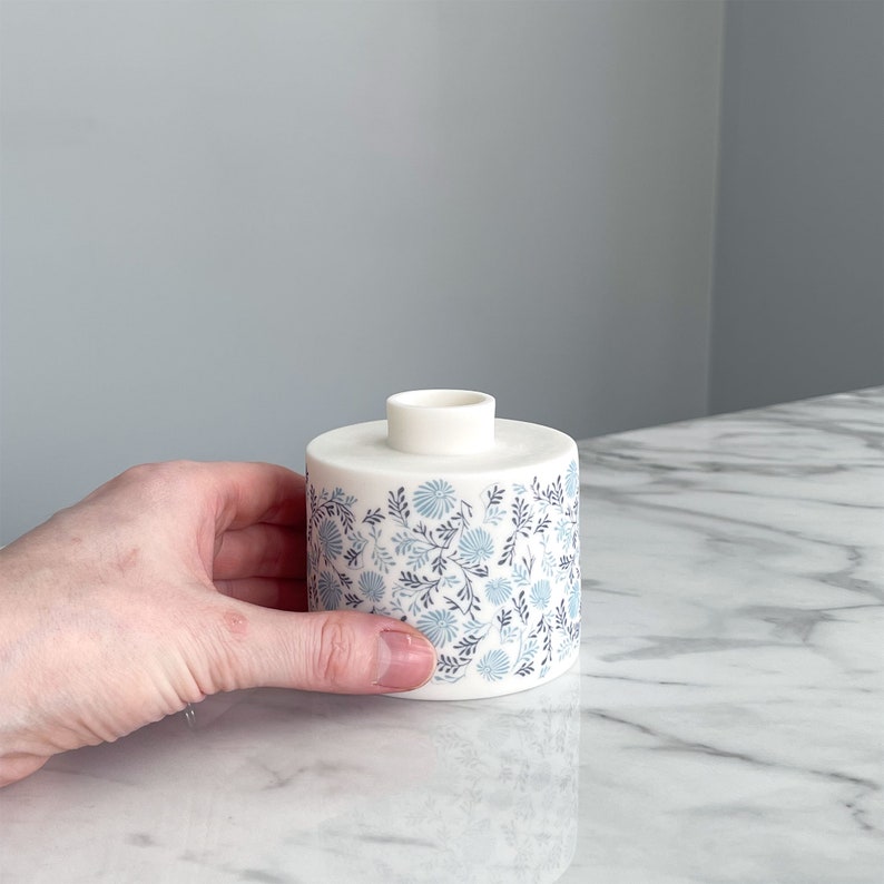 May include: A white ceramic vase with a blue and gray floral pattern. The vase is cylindrical in shape and has a small opening at the top.