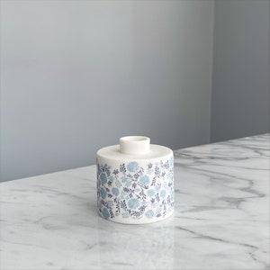 May include: A white ceramic vase with a blue and gray floral pattern. The vase is cylindrical in shape and has a small opening at the top.