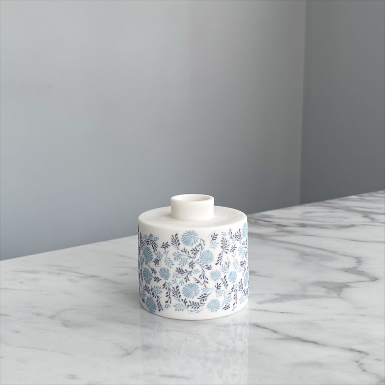 May include: A white ceramic vase with a blue and gray floral pattern. The vase is cylindrical in shape and has a small opening at the top.