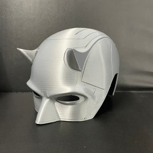 Puede incluir: Un casco gris impreso en 3D con un diseño distintivo, con cuernos puntiagudos y aberturas para los ojos. El casco tiene un acabado liso y mate y parece ser una réplica de una máscara de superhéroe. El tema general es cosplay o disfraz.