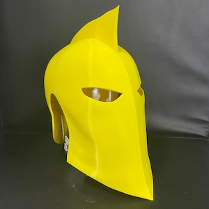 May include: A bright yellow helmet with a pointed top and eye openings. The helmet has a smooth, matte finish and a distinctive, angular design. This cosplay accessory is likely 3D-printed.