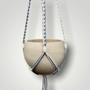May include: A beige ceramic hanging planter with a braided white and blue macrame hanger. The planter has a rounded shape and a tassel at the bottom. The macrame design features a geometric pattern.
