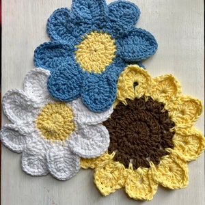 Peut inclure: Trois fleurs en crochet, une bleue, une blanche et une jaune avec un centre brun. Les fleurs sont disposées en grappe.