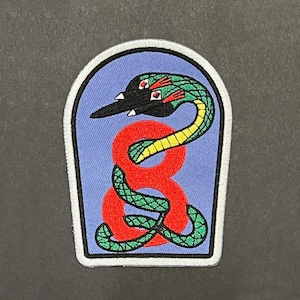 May include: An embroidered patch with a light blue background featuring a large red number eight intertwined with a green snake. The snake has a black head with red and white accents. The patch has a white border and a black inner outline.
