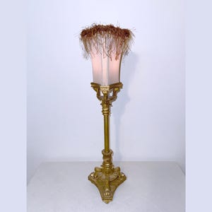 May include: A tall table lamp featuring a gold-toned metal base with an ornate, tripod-style stand. The rectangular shade is topped with a thick, textured fringe of brown fibers, creating a distinct contrast against the metallic structure.