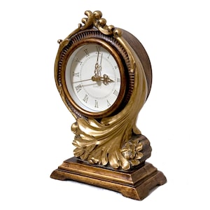 May include: A table clock with a round white face featuring black Roman numerals and gold-colored hands. The clock is encased in a gold and brown frame with ornate leaf-like carvings and a rectangular base. The word QUARTZ is printed on the face.