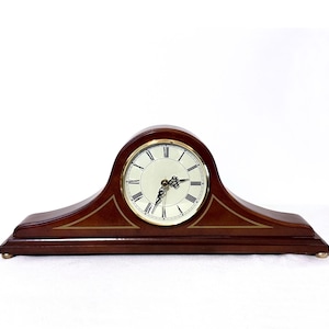 May include: A mantel clock with a dark brown wooden arched frame and a gold-toned bezel. The clock face is off-white with black Roman numerals and ornate black hands. The base features gold-toned line accents and small, rounded gold-toned feet.