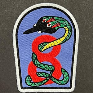 May include: Embroidered patch featuring a stylized snake over a red figure eight. The snake is green, yellow, and black, with red accents. The background is blue, and the patch has a white border. This patch is suitable for jackets, bags, or other fabric items.